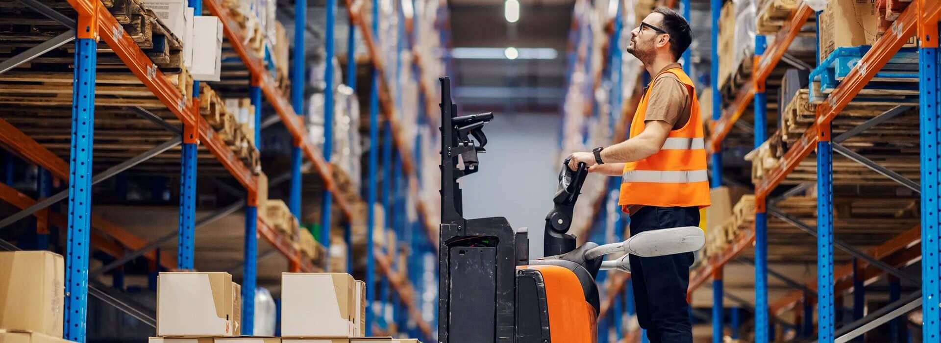 A worker in an orange safety vest operates a forklift in a warehouse, surrounded by shelves filled with boxes.