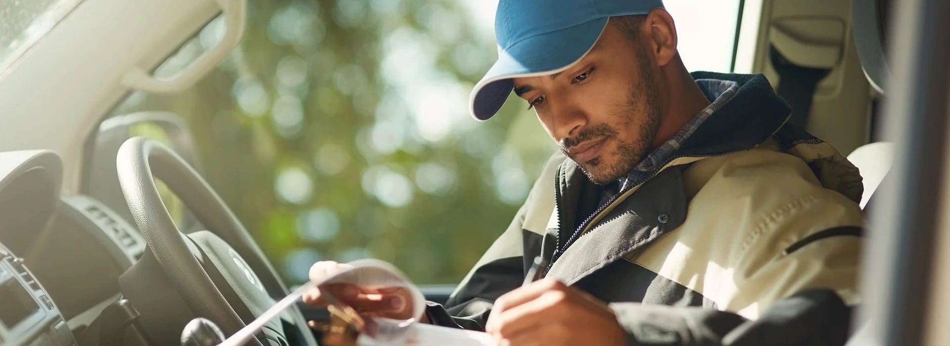 A person wearing a blue cap and jacket sits in a vehicle, looking at a document with a focused expression.