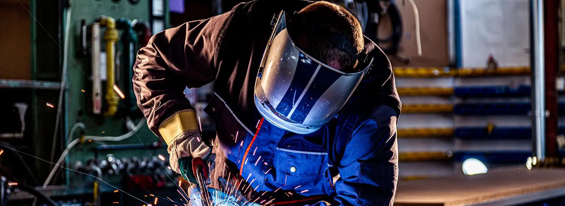 Person in protective gear welding metal in a workshop, with sparks visible.